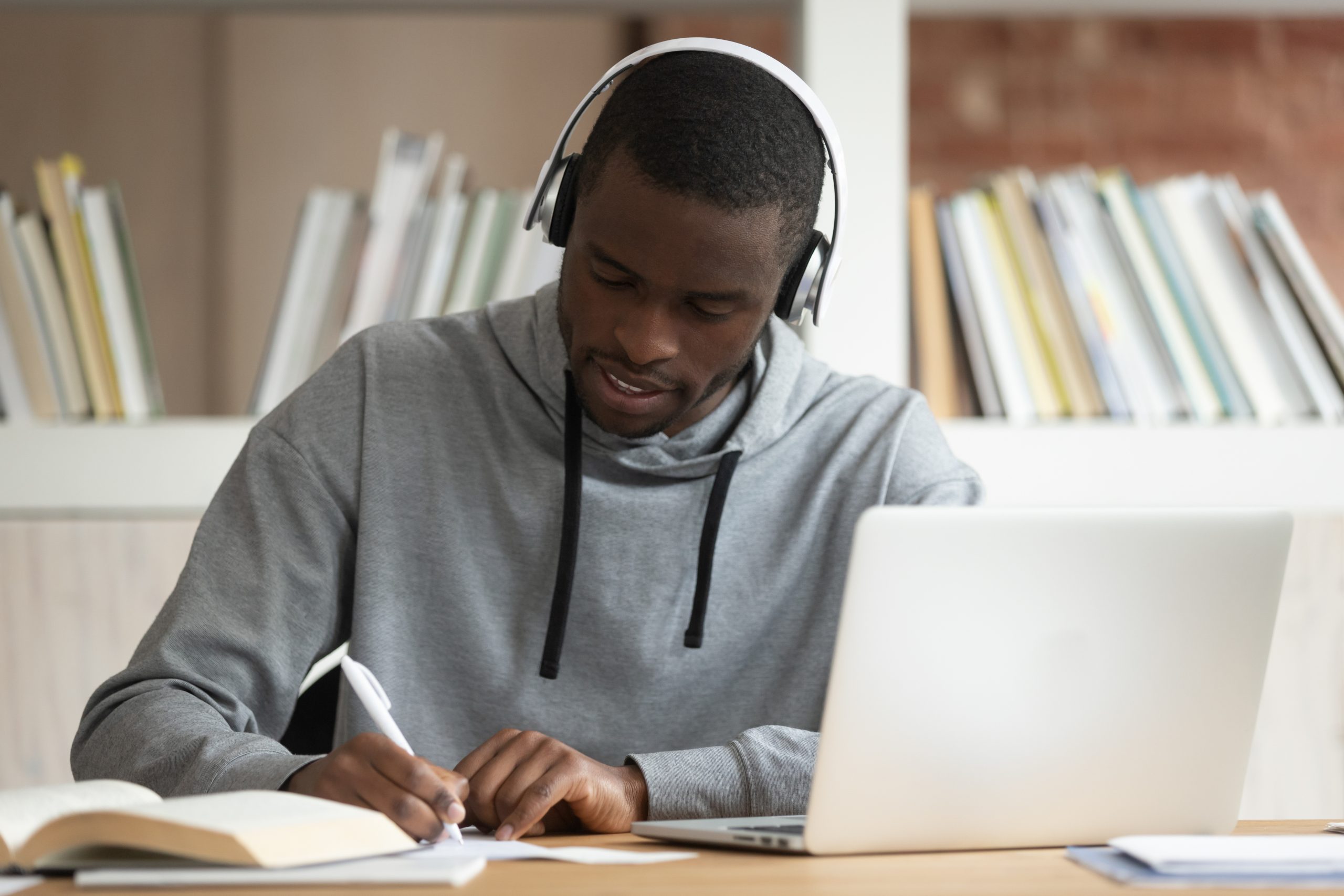 Focused young african american male student sitting at table with computer, wearing bluetooth earphones, busy with study, listening to educational online lecture or enjoying favorite music tracks.